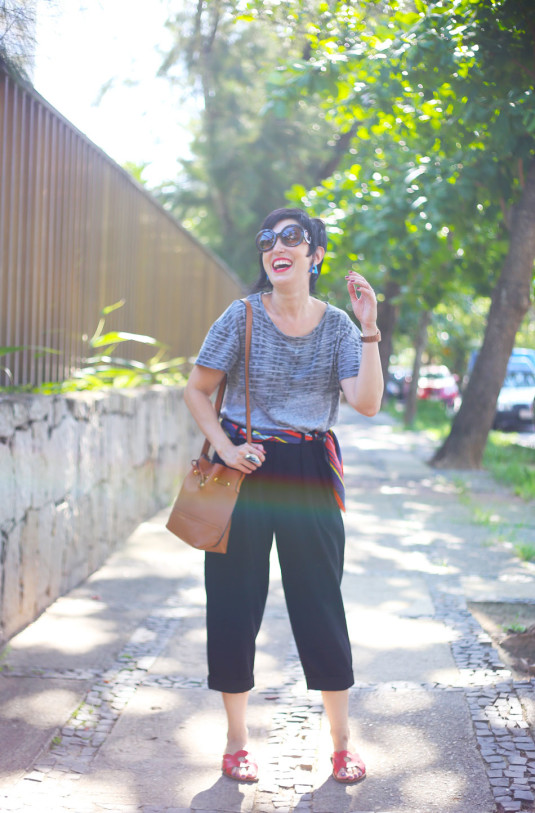 calça alfaiataria curta preta, lenço vermelho, azul escuro e claro e amarelo na cintura, sandália tipo chinelo vermelho, camiseta cinza e brincos triângulo de madeira com detalhes azuis