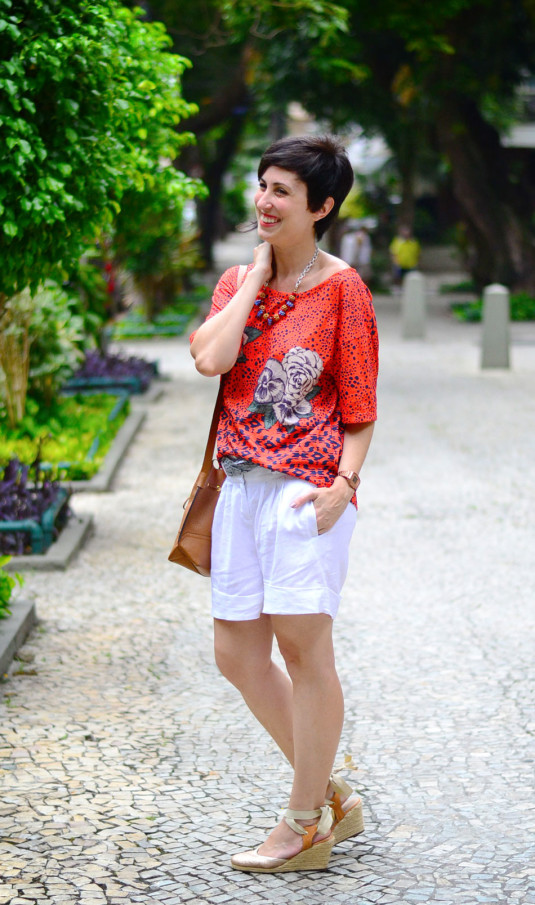 blusa laranja com pintas azuis e rosas brancos e folhas verdes, colar azul, vermelho e dourado, bermuda de linho branca com lenço cinza, espadrilhe dourada e bolsa saco caramelo