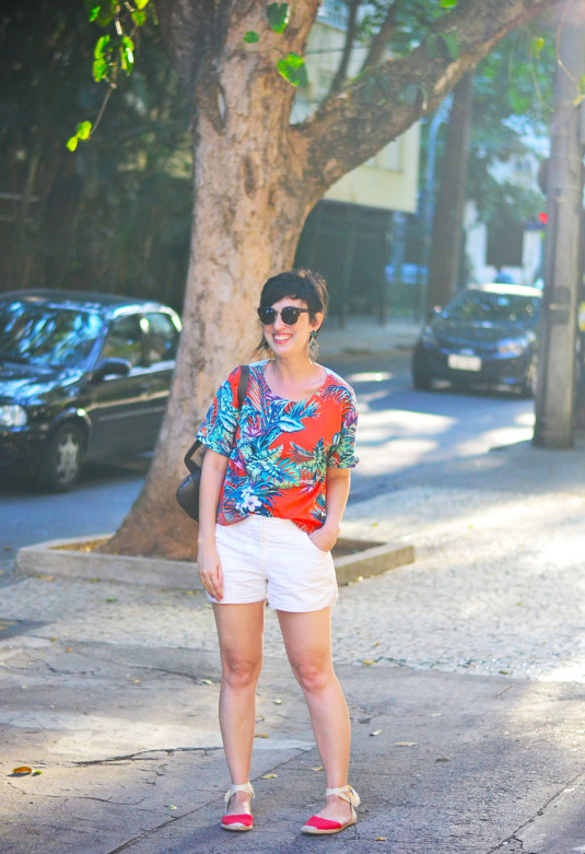 blusa laranja estampada de folhagens tipo palmeiras verdes, azuladas e roxas, short branco, brinco de franjas e triângulos verdes, espadrilhe baixa vermelha