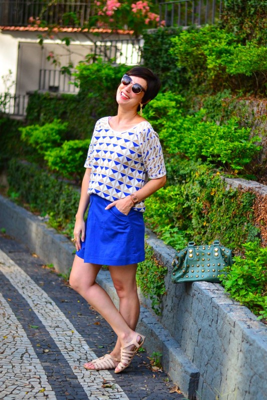 blusa de manga de linha branca com triângulos azuis de dourados, saia azul trapézio, sandália de tiras nude, bolsa verde de tachas