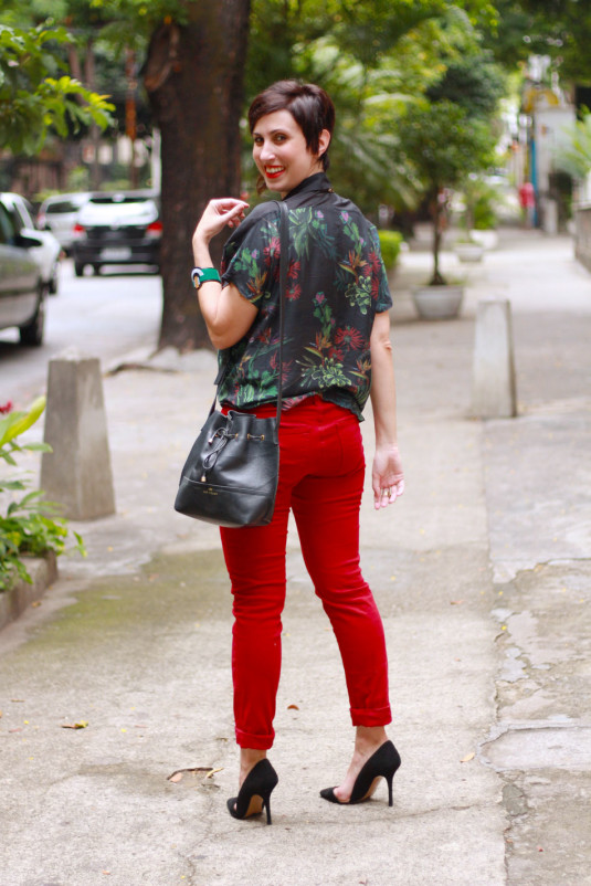 calca-vermelha skinny, scarpin vazado lateral preto, blusa com gola camisa mangas curtas preta com estampa de folhagens verdes e vermelhas, colar triangulos dourados sob gola e bolsa saco preta