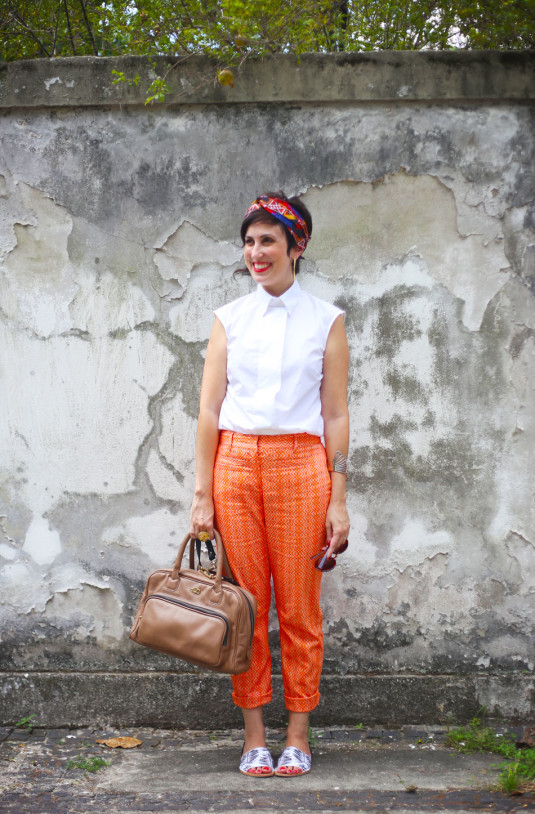 calça laranja com motivos geométricos, camisa branca sem mangas com gola abotoada, len~co vermelho, azul e amarelo como faixa na cabeçca, sandália rasteira com faixa grossa na frente branca e estampa de folhagens de palmeiras pretas, bolsa estilo maleta marrom clara