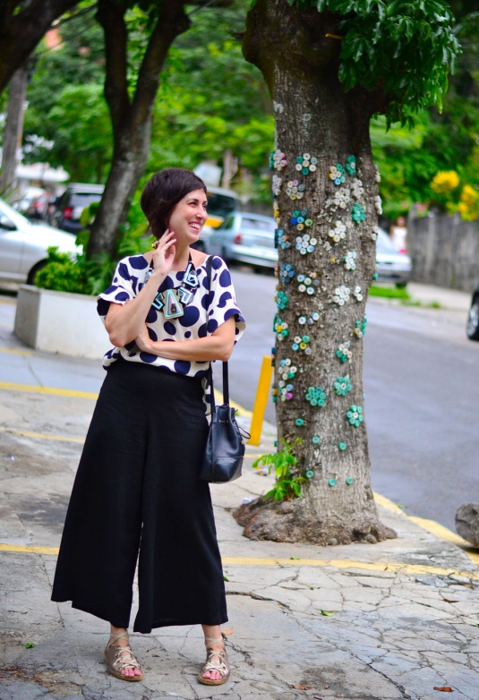 blusa de mangas curtas branca com bolas azuis marinho, calça pantalona curta preta, sandália de amarrar dourada e solado de corda, colar gigante geométrico preto, rosa e azul