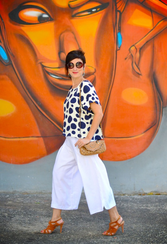 pantalona ampla branca curta, sandália de tiras trançadas caramelo, blusa soltinha de mangas curtas branca com estampas de bolas azul escuras e bolsa pequena de tachas caramelo clara